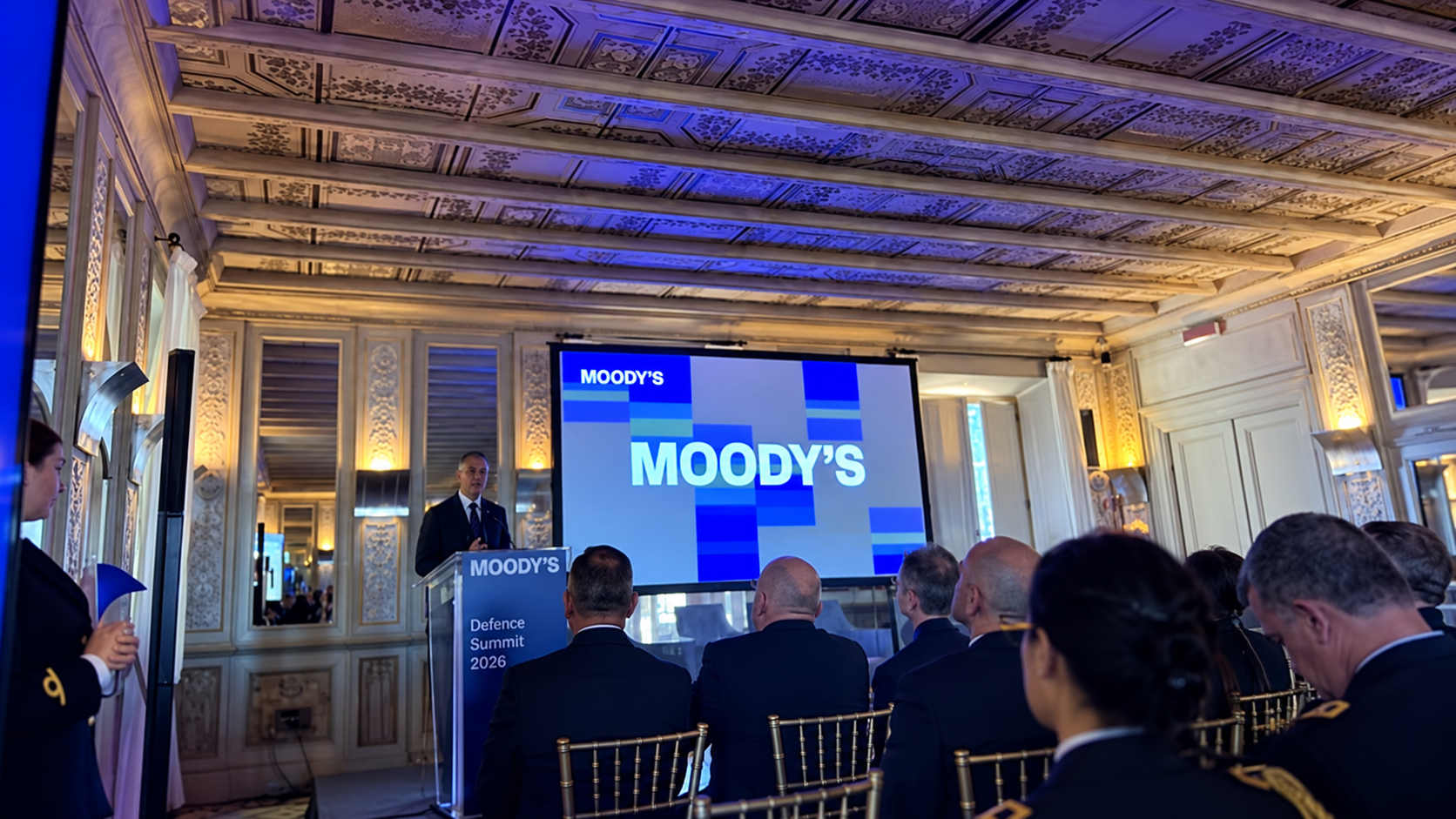 Elaborazione editoriale del Moody’s Defence Summit 2026: sala conferenze con pubblico, relatore al leggio e schermo Moody’s